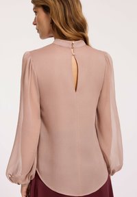 Kobieta o jasnobrązowych włosach nosi przezroczystą, długą bluzkę w jasnoróżowym kolorze z dekoltem w kształcie łezki i perłowymi guzikami przy szyi.