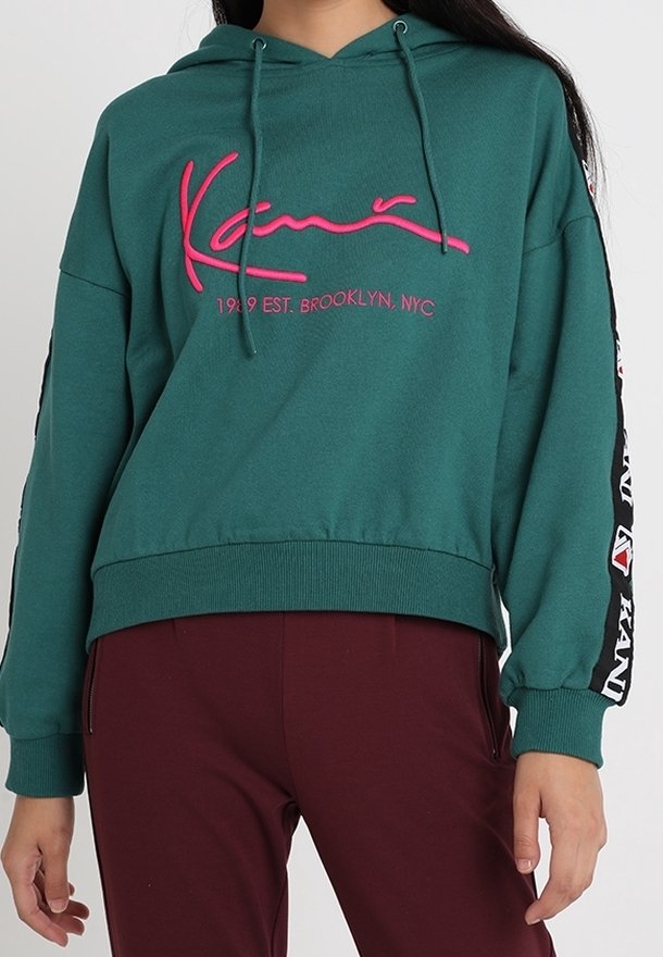 Sweat à capuche turquoise avec texte rose "Kani 1989 EST. BROOKLYN, NYC", cordons noirs et manches à rayures latérales, associé à un pantalon bordeaux.