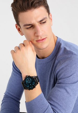 Junger Mann im blauen Pullover sieht auf seine schwarze Chronographenuhr am Handgelenk vor einem schlichten hellen Hintergrund.