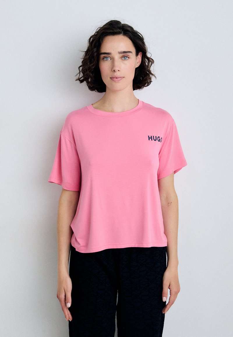 Rosa t-shirt med kort ärm och avslappnad passform, med en liten svart "HUG"-logga på vänster bröst. Mjuk, jämn tygtextur.