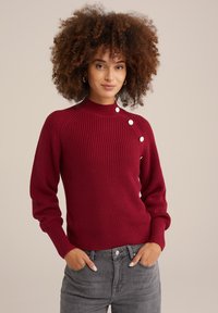 Bordo megztinis su raukšlėmis, aukštu kaklu, raglan stiliumi ir dekoratyvinėmis baltomis mygtukais ant peties. Suderintas su pilkais džinsais.