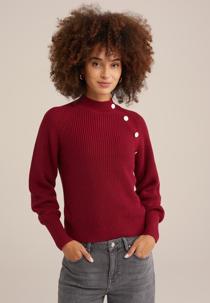 Bordo megztinis su raukšlėmis, aukštu kaklu, raglan stiliumi ir dekoratyvinėmis baltomis mygtukais ant peties. Suderintas su pilkais džinsais.