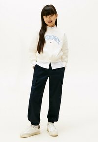 Felpa bianca con "TOMMY" in lettere blu, indossata sopra una camicia bianca. Pantaloni cargo blu scuro con tasche, abbinati a sneakers bianche.