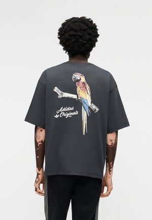Mann med mørkt krøllete hår og vitiligo, iført mørk, oversized t-skjorte med en fargerik papegøye sittende på en gren og tekst "Adidas Originals" på baksiden.