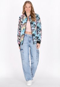 Giacca bomber floreale in colori pastello con dettagli neri, abbinata a pantaloni cargo azzurri chiari e sneakers bianche. Texture del tessuto liscia.