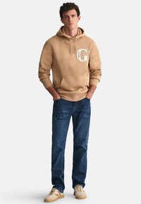 Felpa color marrone chiaro con tasca a marsupio, con logo bianco a forma di "G". Abbinata a jeans blu scuro e sneaker beige.