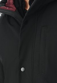 Czarny płaszcz o matowym wykończeniu, z bordową podszewką, z guzikami z przodu i eleganckim krojem. Posiada też szczegóły w postaci bocznych kieszeni.