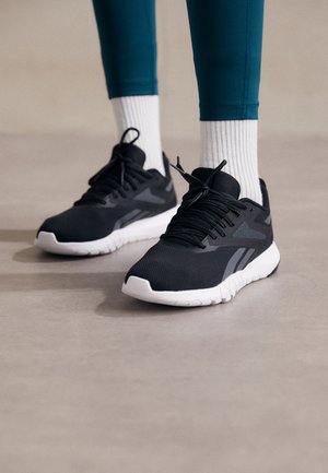 Zapatillas deportivas negras con una parte superior de malla texturizada, suela blanca y acentos grises. Llevadas con calcetas blancas de canalé y leggings en color azul verdoso.