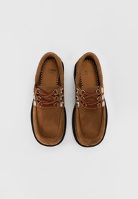 Chaussures en daim marron avec une forme arrondie, des lacets rouges et des éléments décoratifs en métal argenté. La semelle est en caoutchouc noir, avec un motif texturé.