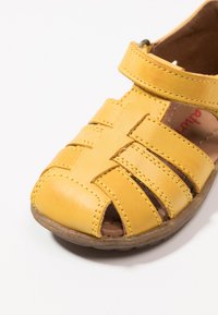 Žluté kožené sandály s překříženými pásky, otevřenou špičkou, hnědou stélkou a strukturovanou gumovou podrážkou. Upravují se pomocí pásku a přezky.