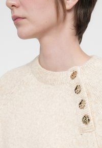 Maglione lavorato a maglia color crema con colletto a costine, caratterizzato da quattro bottoni decorativi in tono oro lungo la spalla per un tocco in più.