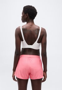 Αθλητική γυναίκα με κοντά μαλλιά που φοράει λευκό αθλητικό σουτιέν και ροζ σορτς Under Armour, στέκεται με την πλάτη προς τα εμπρός σε απλό φόντο.