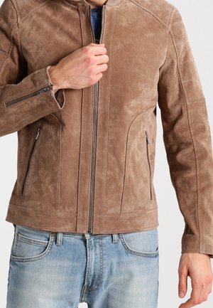 Homme portant une veste en daim marron clair et un jean bleu clair, la main tenant la fermeture éclair de la veste au niveau de la poitrine.