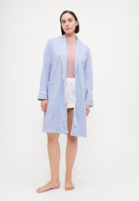 Lättviktig bomullsrock med blå och vita vertikala ränder, långa ärmar, öppen front och två sidofickor. Buren över en rosa topp och shorts.