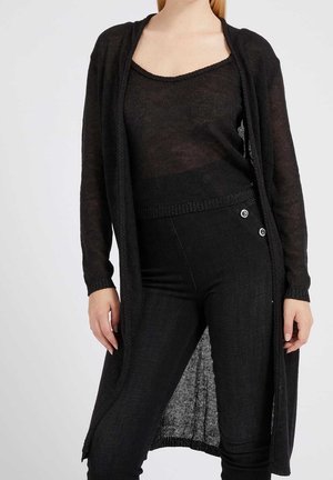 Frau trägt eine durchsichtige schwarze langärmelige Strickjacke, ein passendes ärmelloses Oberteil und hoch taillierte schwarze Hosen mit seitlichen Knöpfen.