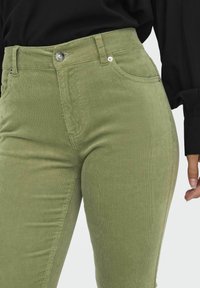 Pantalons en velours côtelé verts avec une taille haute, présentant une fermeture à glissière et un bouton, des poches avant et une finition lisse et texturée.