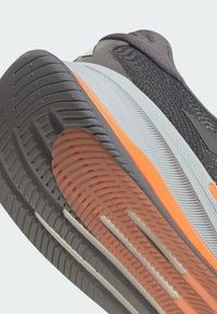 Scarpa da ginnastica grigia con una parte superiore testurizzata, un accento arancione sulla suola intermedia e una suola in gomma con motivo per una migliore aderenza.