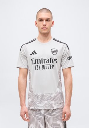 Homme portant un maillot de football gris d'Arsenal avec le logo du sponsor « Emirates Fly Better » et un short à motif abstrait, debout devant un fond uni.