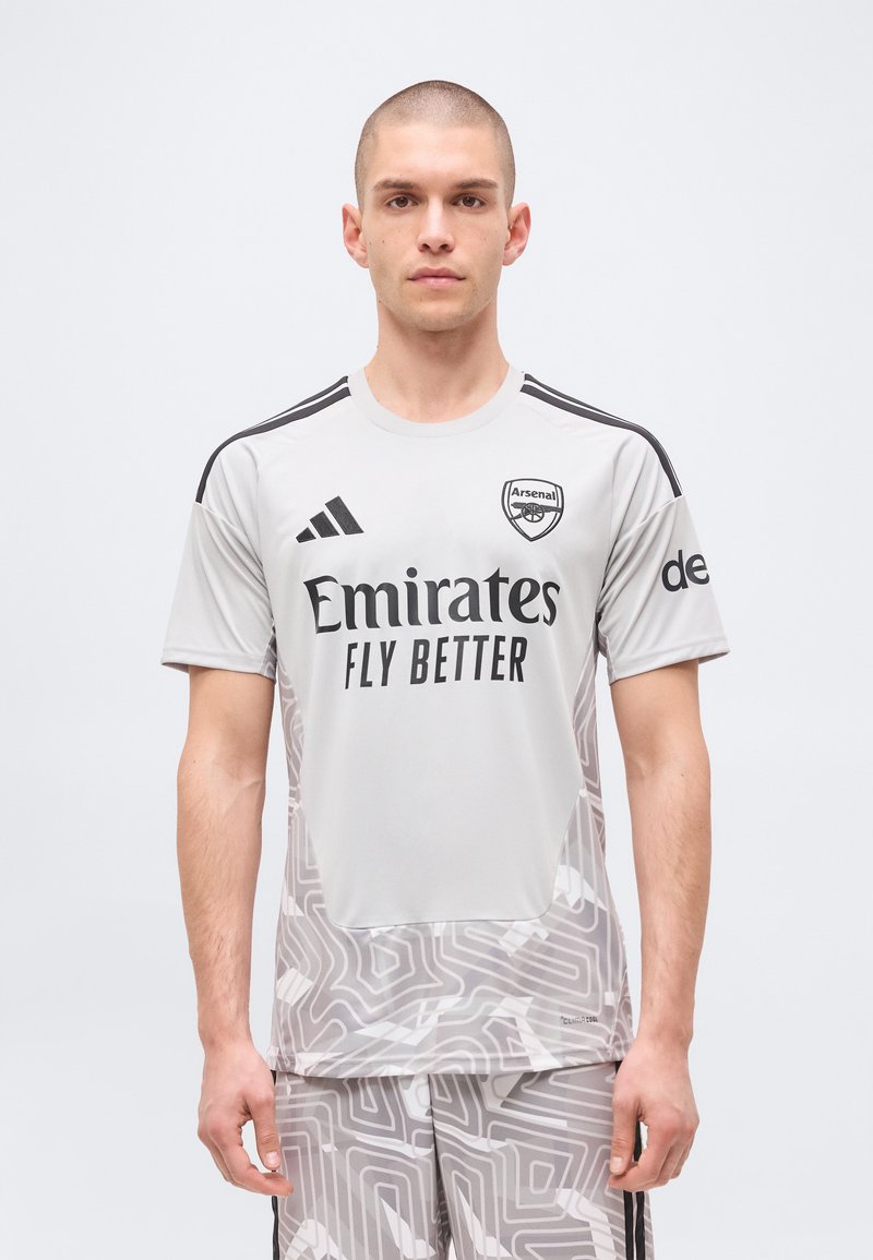 Uomo che indossa la maglia da calcio grigia dell'Arsenal con il logo dello sponsor "Emirates Fly Better" e pantaloncini dal motivo astratto, in piedi davanti a uno sfondo semplice.