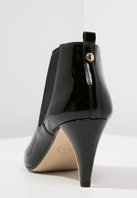 Bottine en cuir verni noir avec un talon effilé, dotée de panneaux latéraux élastiques et d'une touche dorée à l'arrière. Texture lisse.