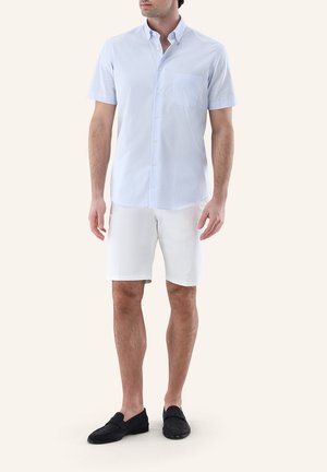 Mann trägt hellblaues kurzärmeliges Hemd mit Knöpfen, weiße knielange Shorts und schwarze Slipper, steht vor einfarbigem Hintergrund.