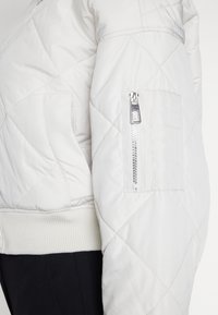 Vadderad vit jacka med ribbat avslut, utrustad med en sidofick med dragkedja och en mjuk textur. Designen inkluderar framträdande diamantmönster.