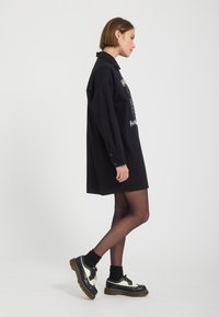 Jeune femme aux cheveux courts portant une robe noire ample à manches longues, des collants noirs transparents et des chaussures à plateforme noires et blanches.