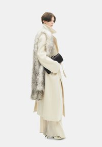 Lange crèmekleurige jas met een bontsjaal, zwarte gestructureerde clutch en lichte schoenen, die een elegante, minimalistische uitstraling tonen.