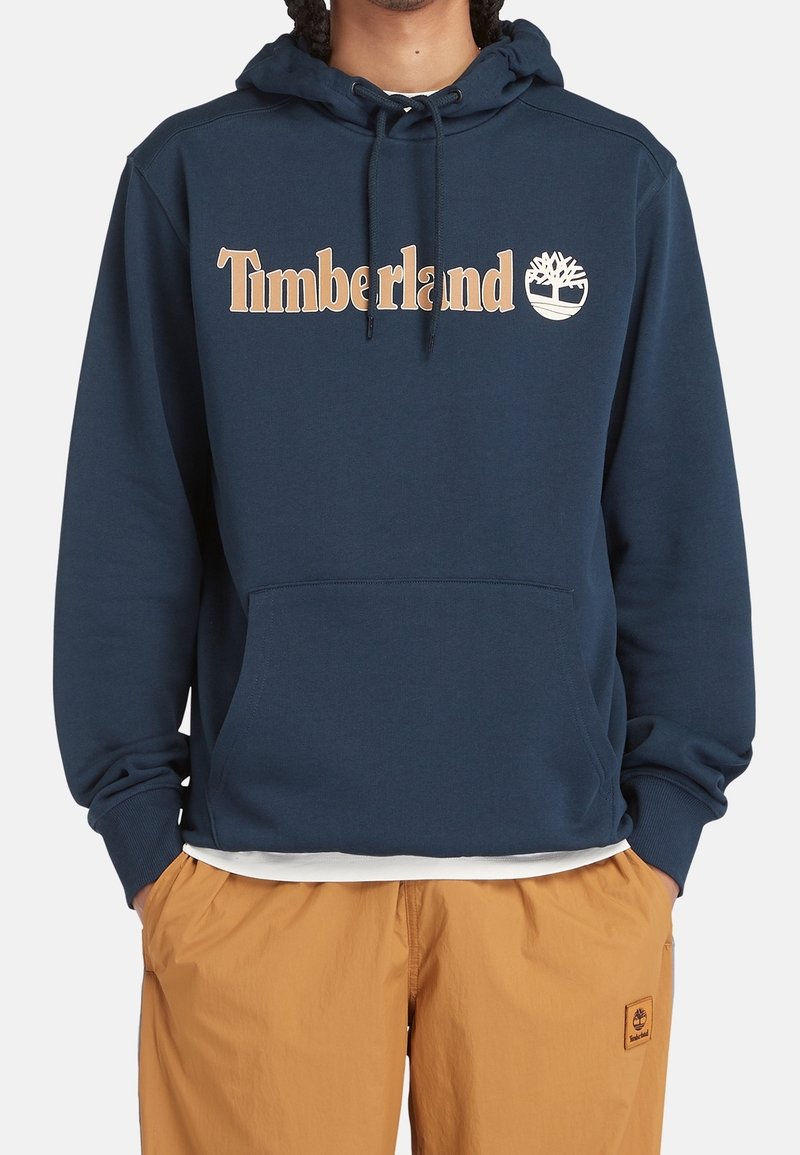 Personne portant un sweat à capuche Timberland bleu marine avec logo beige et pantalon beige avec patch Timberland, mains dans les poches, debout devant un fond blanc.