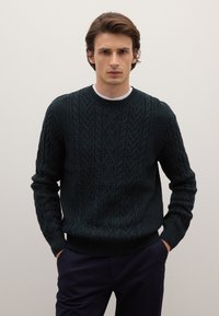 Dunkelgrüner Grobstrickpullover mit Rundhalsausschnitt, strukturierten Mustern sowie gerippten Bündchen und Saum. Wird über einem weißen Hemd getragen.