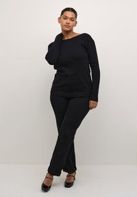 Černý top s dlouhým rukávem a černé slim-fit kalhoty. Top má široký výstřih; obě součásti jsou vyrobeny z hladkého materiálu. Černé lesklé boty dotvářejí vzhled.