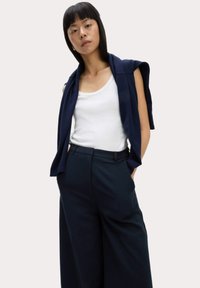 Navy vida-byxor kombinerat med en vit linne och en marinblå stickad tröja över axlarna. Slät textur, skräddarsydd passform.