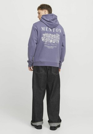 Lilla hættetrøje med et grafisk motiv af Menton på ryggen, der har hvid tekst og design. Parret med løstsiddende mørke jeans.