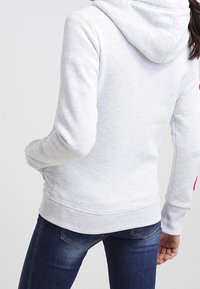 Sudadera con capucha gris de textura suave, con un bolsillo tipo canguro y puños acanalados, combinada con jeans azules. Detalles en las mangas.