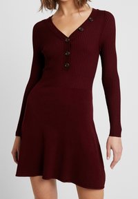 Abito bordeaux a coste con maniche lunghe, scollatura a V, cinque bottoni decorativi e gonna svasata. Tessuto morbido e corpetto aderente.