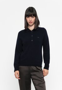 Maglione lavorato a maglia blu navy con colletto polo, tre bottoni e polsini a coste. Tessuto leggero con una texture liscia. Logo sul petto.