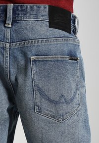 Bakvy av blå denimjeans med sydd fickdesign och svarta märkeslappar, som bärs med en röd topp.