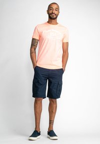 Kratkoročanski koralni majica s kratkimi rokavi in belim tiskom, v kombinaciji z mornarsko modrimi kratkimi hlačami s stranskimi žepi in mornarsko modrimi supergami brez vezalk.