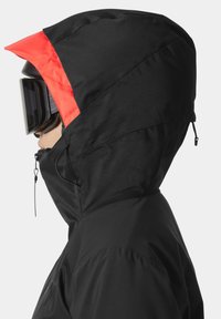 Czarna kurtka narciarska z czerwonym akcentem na kapturze, teksturowana tkanina, regulowane sznurki oraz wysoka stójka zapewniająca dodatkową ochronę.