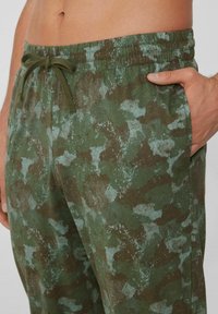 Joggers de camuflaje verde fabricados en un tejido suave, que cuentan con una cinturilla elástica, cordón ajustable y bolsillos laterales con un patrón texturizado.