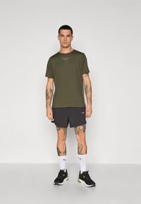 Olive green short-sleeve t-shirt with logo, paired with black shorts. Model wears black athletic shoes and white socks with logo.