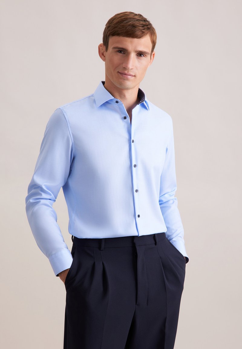 Camicia azzurra chiara con colletto strutturato e bottoni scuri, abbinata a pantaloni scuri e su misura. Tessuto liscio, design aderente.