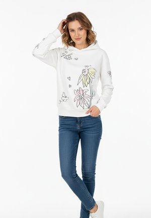 Junge Frau mit schulterlangem Haar, die einen weißen Kapuzenpullover mit Blumen- und Herzmotiven, blaue Jeans und weiße Turnschuhe trägt und steht.