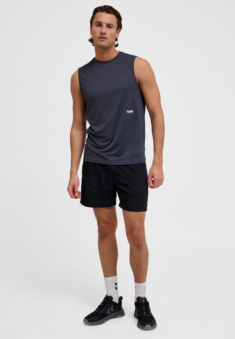 Graues ärmelloses Sportoberteil mit Logo, kombiniert mit schwarzen Shorts. Model trägt schwarze Sneakers und weiße Socken mit schwarzen Akzenten.