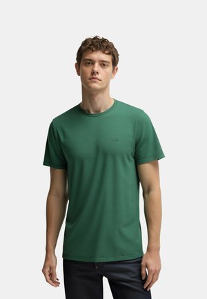 Ragazzo giovane con capelli ricci che indossa una maglietta verde semplice a maniche corte e pantaloni scuri, in piedi davanti a uno sfondo bianco.