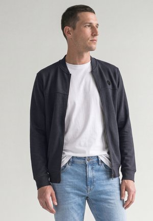 Jonge man staand met een zwarte jas met rits, wit T-shirt en lichtblauwe spijkerbroek tegen een egale lichte achtergrond.