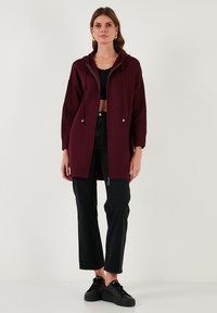 Moteris stovi apsistrenčiusi ilgu bordo spalvos užtraukiamu džemperiu, juoda trumpa palaidine, juodomis kelnėmis, juodais sportbačiais ir geometriniais auksiniais auskarais ant vienodo fono.