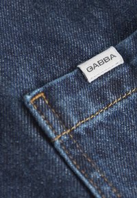 Sötétkék lemosott farmeranyag, látható textúrával és varrással. Egy kis fehér címke, amelyen az áll, hogy "GABBA", a zsebszegélyhez van erősítve.