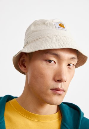 Beige bucket hat with a round crown, stitched brim, and a square patch with an orange logo. Fabric appears textured and casual.