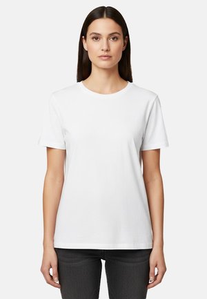 Donna con capelli lisci e scuri che indossa una maglietta bianca a maniche corte e pantaloni scuri, in piedi davanti a uno sfondo chiaro.